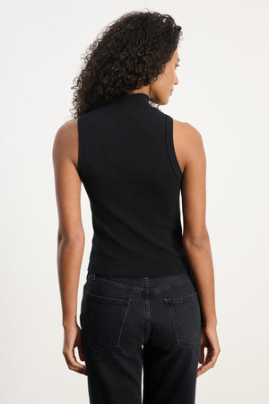 A woman faces away, wearing the CEAN TANK TOP by Velvet by Graham & Spencer—a cropped, ribbed cotton tank—paired with black jeans against a plain, light background.