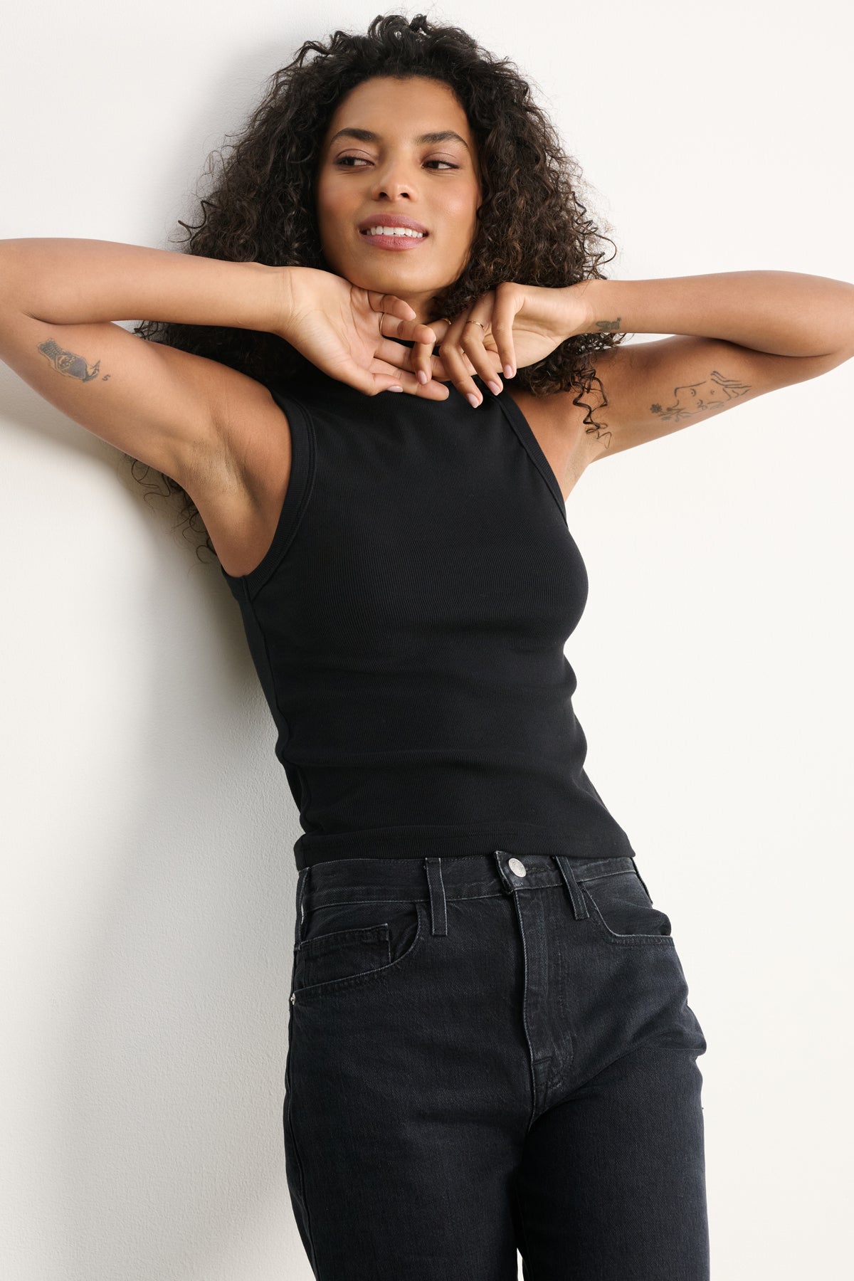 A woman with curly hair wears the Velvet by Graham & Spencer CEAN TANK TOP—a cropped, ribbed cotton tank—paired with black jeans, standing against a white wall with arms raised and a slight smile.