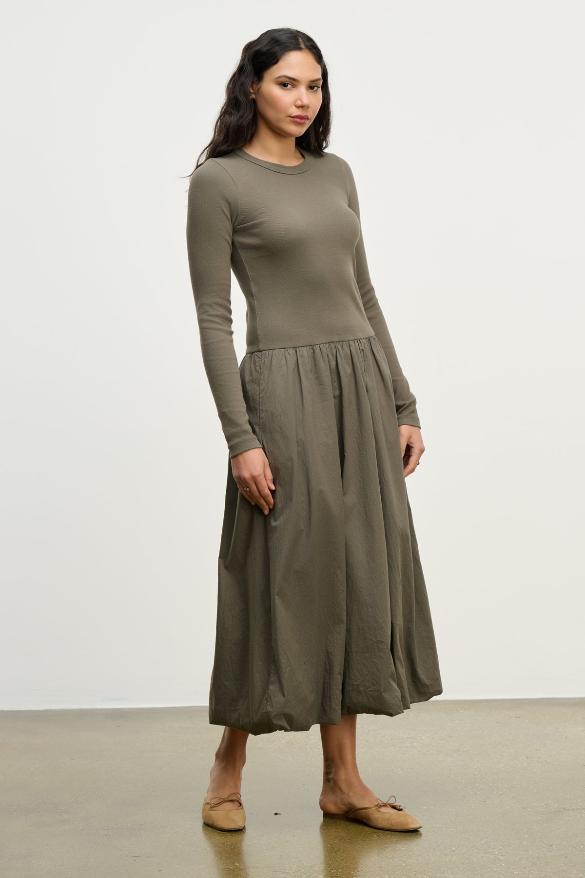 A woman stands against a plain background, wearing the DOLCE DRESS by Velvet by Graham & Spencer—an olive-green, long-sleeve cotton poplin dress with a gathered skirt—paired with tan flats.