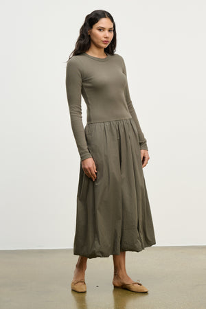 A woman stands against a plain background, wearing the DOLCE DRESS by Velvet by Graham & Spencer—an olive-green, long-sleeve cotton poplin dress with a gathered skirt—paired with tan flats.