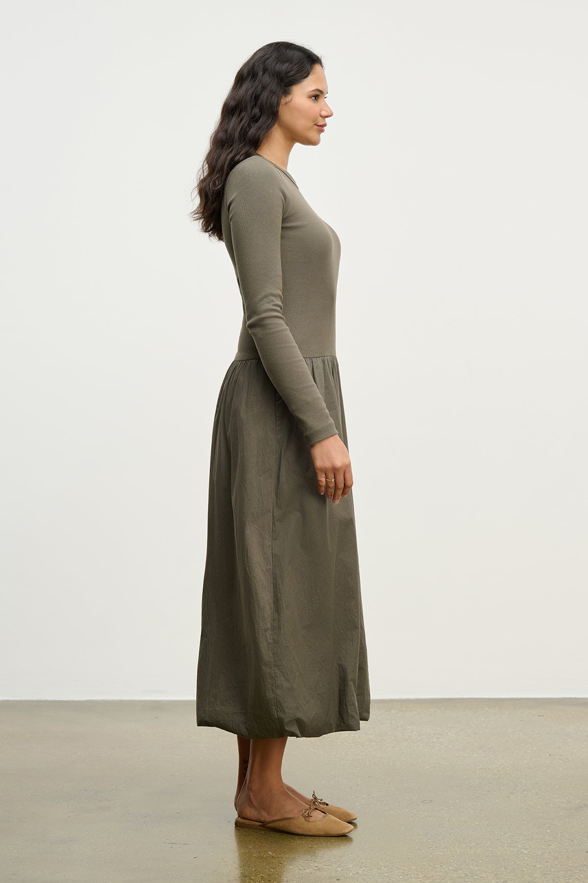 A woman in profile wears the DOLCE DRESS by Velvet by Graham & Spencer, a long-sleeved ribbed cotton dress in olive green, paired with brown flats, against a plain white background.