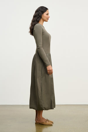 A woman in profile wears the DOLCE DRESS by Velvet by Graham & Spencer, a long-sleeved ribbed cotton dress in olive green, paired with brown flats, against a plain white background.