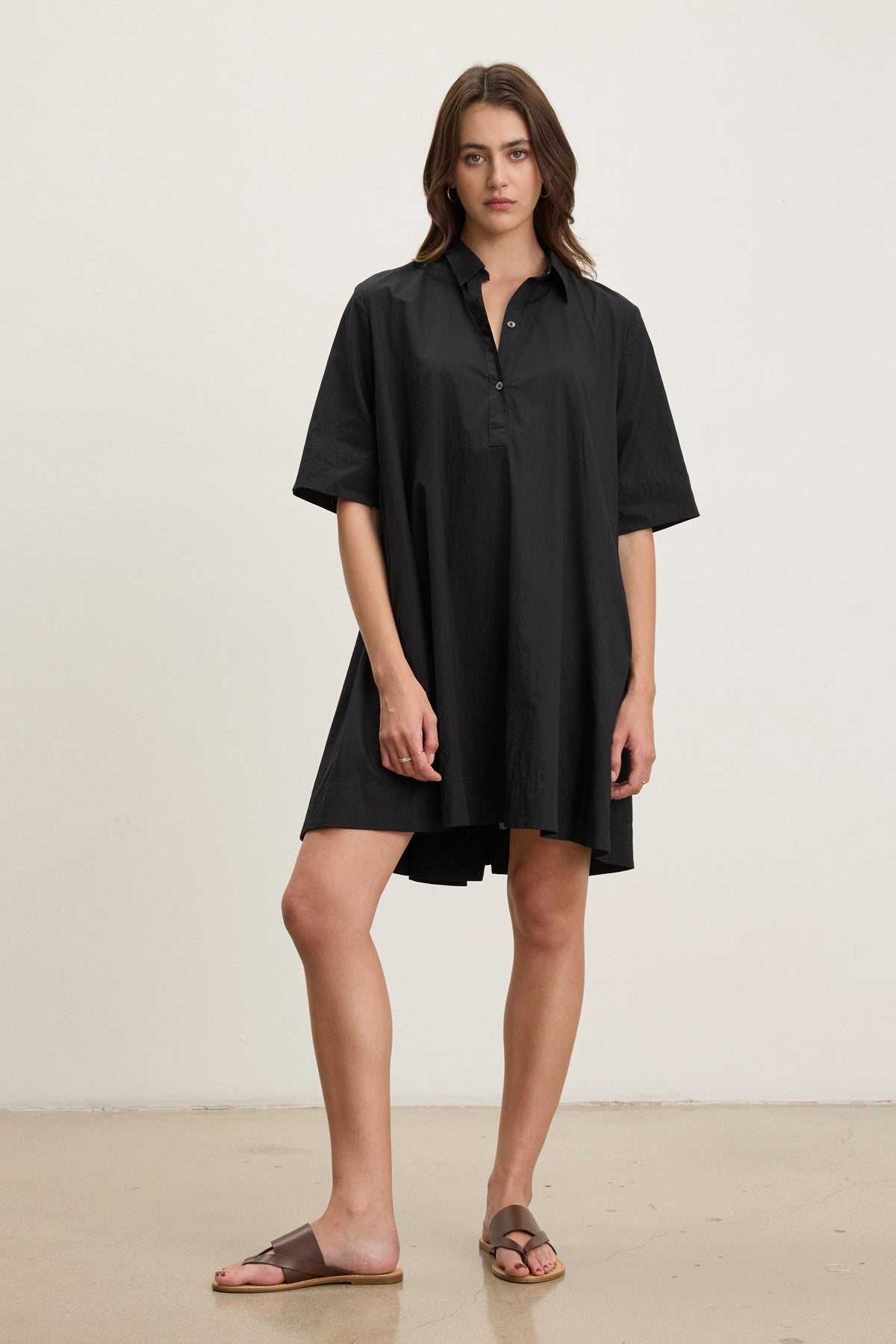 A woman stands indoors against a plain light background, wearing the Velvet by Graham & Spencer DRIANA DRESS—a loose-fitting black cotton poplin dress with half sleeves and side seam pockets—paired with brown slide sandals.-39145480782017