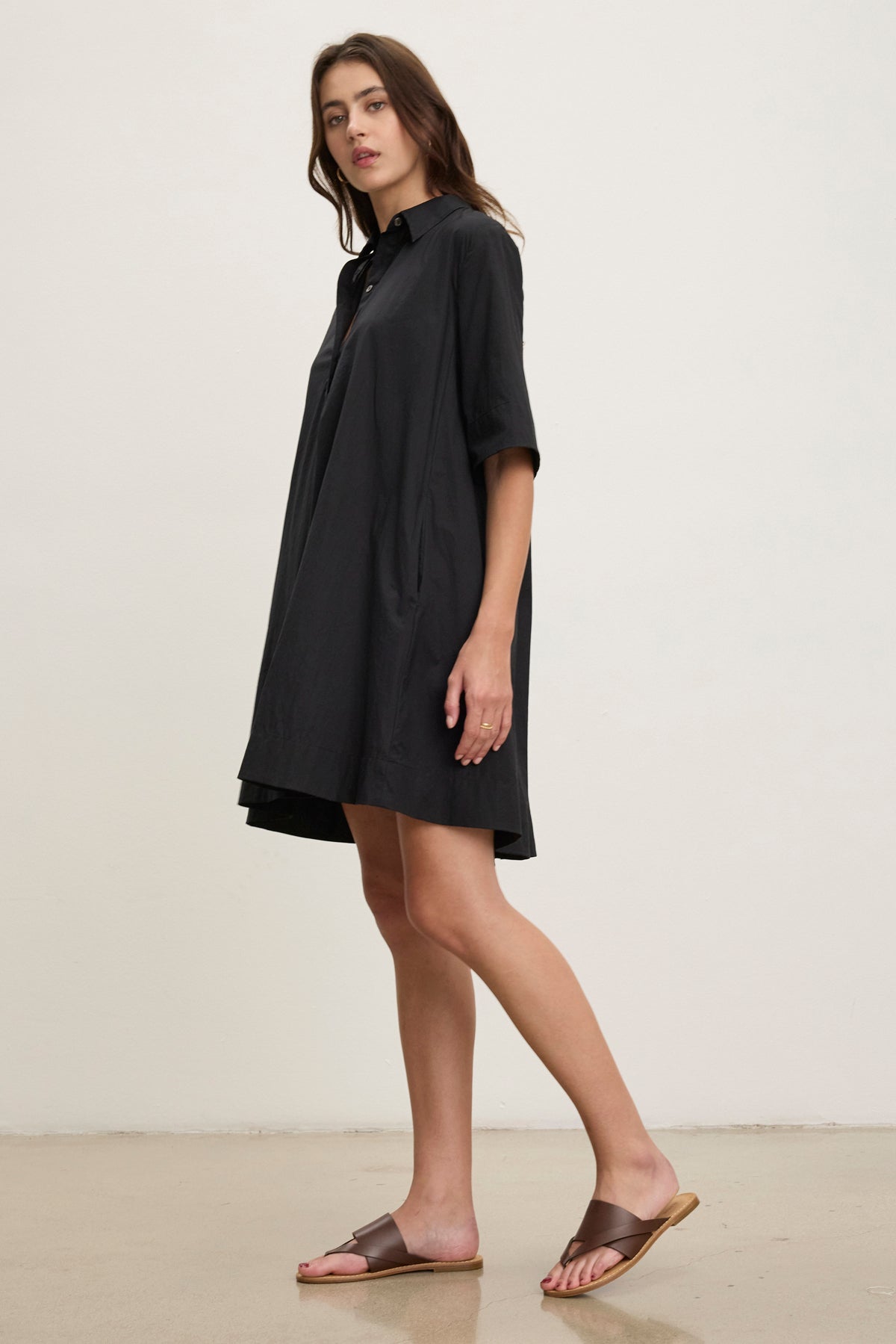A woman stands sideways against a plain, light background wearing the DRIANA DRESS by Velvet by Graham & Spencer—a loose black cotton poplin dress with short sleeves, side seam pockets, and brown slide sandals.
