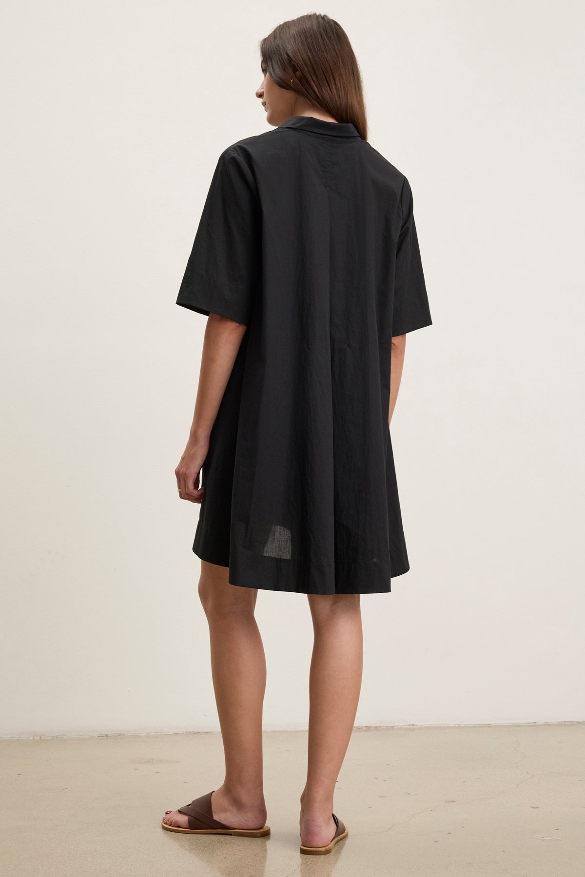 A person stands indoors facing away, wearing the DRIANA DRESS by Velvet by Graham & Spencer—a loose-fitting, short-sleeve black cotton poplin dress with side seam pockets—and brown slide sandals on a light floor.