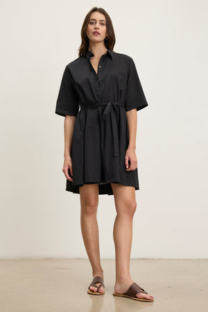 A woman stands against a plain background wearing the DRIANA DRESS by Velvet by Graham & Spencer, a black cotton poplin dress with a detachable belt and short sleeves, paired with brown slide sandals.