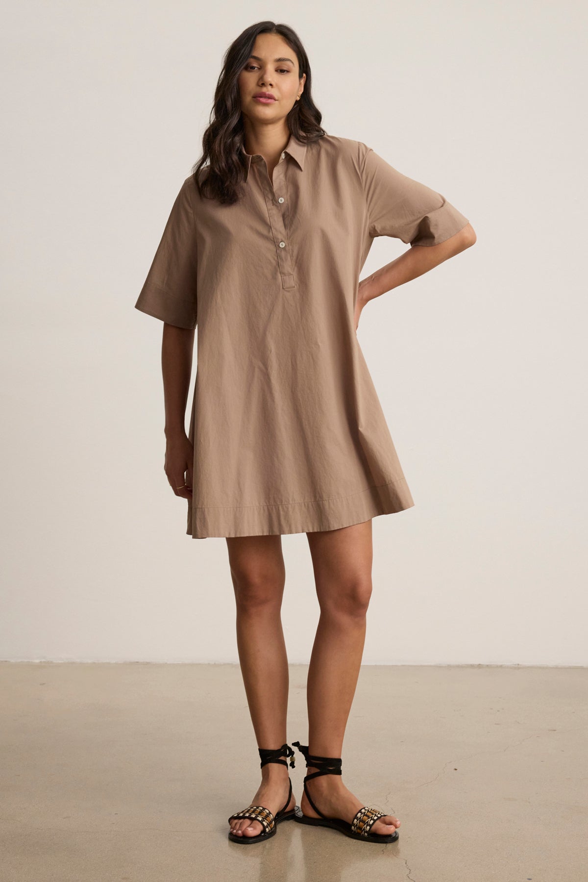 A woman stands indoors wearing the DRIANA DRESS by Velvet by Graham & Spencer—a loose-fitting tan cotton poplin dress with short sleeves and side seam pockets—paired with black sandals. One hand rests on her hip as she maintains a neutral expression.