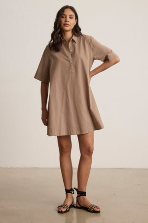 A woman stands indoors wearing the DRIANA DRESS by Velvet by Graham & Spencer—a loose-fitting tan cotton poplin dress with short sleeves and side seam pockets—paired with black sandals. One hand rests on her hip as she maintains a neutral expression.