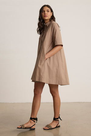 A woman wears the DRIANA DRESS by Velvet by Graham & Spencer—a loose, tan cotton poplin style with front and side seam pockets—paired with black sandals, set against a plain background.