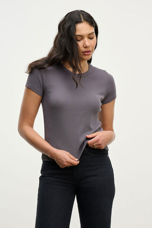 A woman with long, wavy hair wears the EDDIE TEE by Velvet by Graham & Spencer—a fitted, cropped crewneck tee—paired with black jeans as she looks down and holds the hem of her shirt against a plain background.