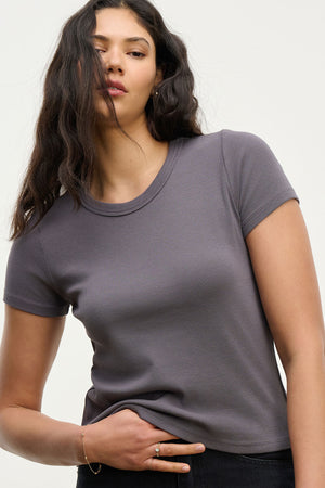 A woman with long dark hair wears the Velvet by Graham & Spencer EDDIE TEE, a fitted cropped crew-neck tee, paired with dark pants and standing against a plain light background.