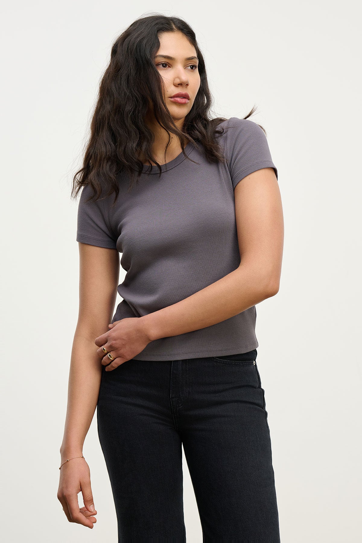 A woman with long dark hair wears the EDDIE TEE by Velvet by Graham & Spencer in gray, paired with black pants, standing against a plain light background.