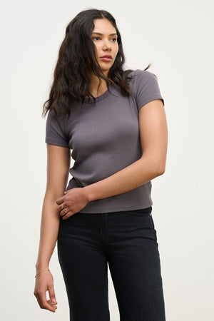 A woman with long dark hair wears the EDDIE TEE by Velvet by Graham & Spencer in gray, paired with black pants, standing against a plain light background.