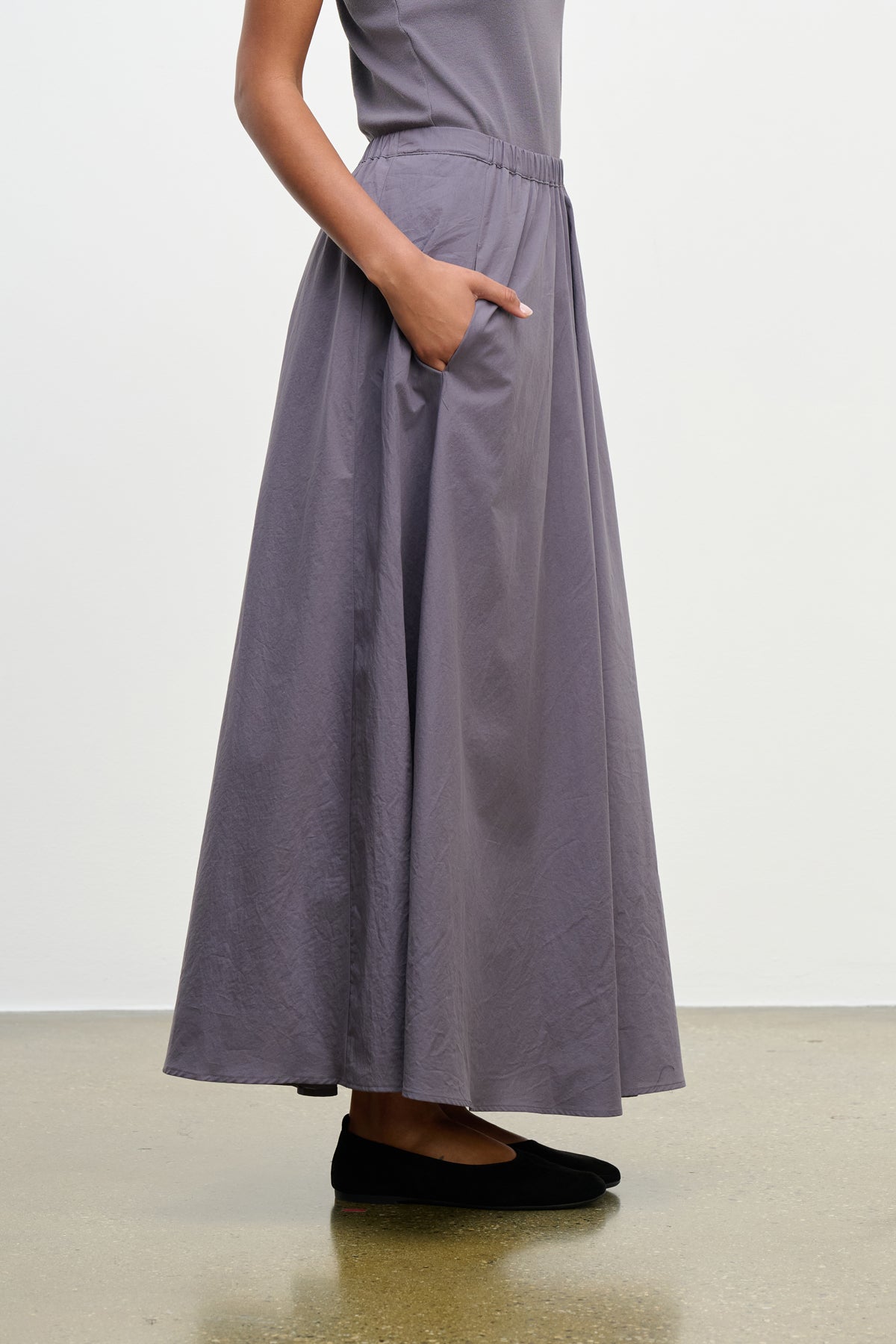 Model wearing the FILLA SKIRT by Velvet by Graham & Spencer—a grey, ankle-length elastic waist skirt with pockets—paired with a grey top and black flats, standing on a polished floor against a plain white background.