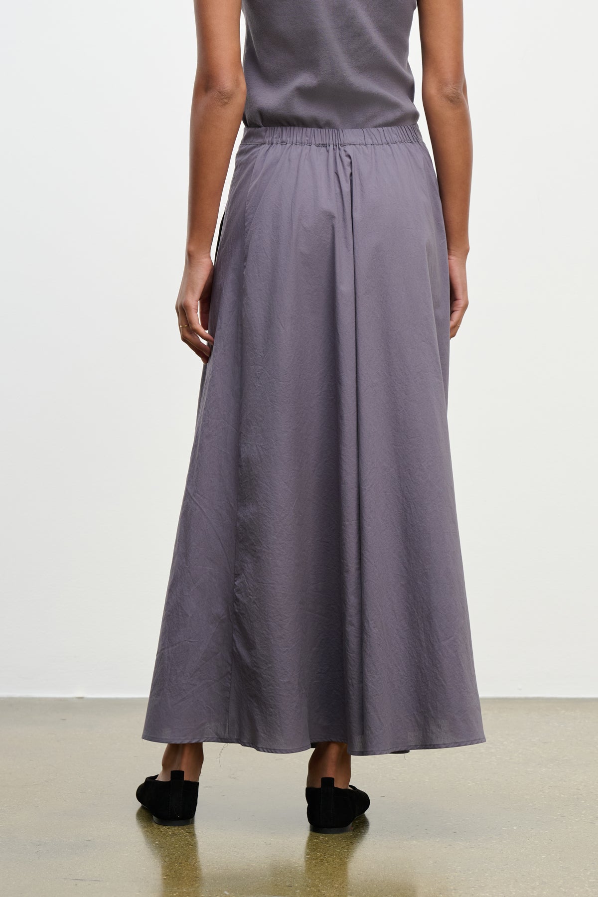A person wearing a gray sleeveless top and the FILLA SKIRT by Velvet by Graham & Spencer, a long flowy cotton poplin skirt with an elastic waist, stands facing away from the camera on a polished floor.