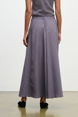 A person wearing a gray sleeveless top and the FILLA SKIRT by Velvet by Graham & Spencer, a long flowy cotton poplin skirt with an elastic waist, stands facing away from the camera on a polished floor.