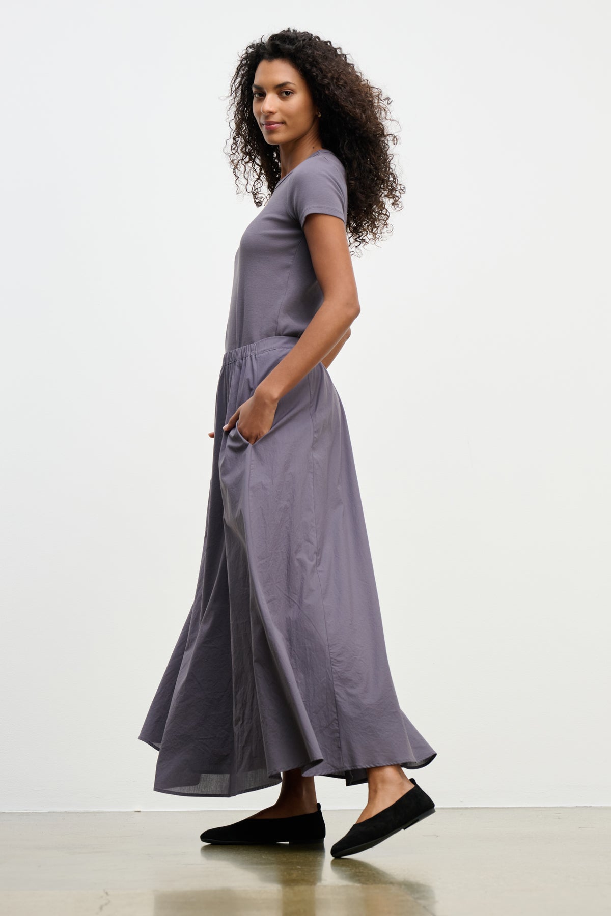A woman stands sideways with hands in the pockets of the gray FILLA SKIRT by Velvet by Graham & Spencer, wearing black flat shoes against a plain background.
