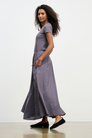 A woman stands sideways with hands in the pockets of the gray FILLA SKIRT by Velvet by Graham & Spencer, wearing black flat shoes against a plain background.
