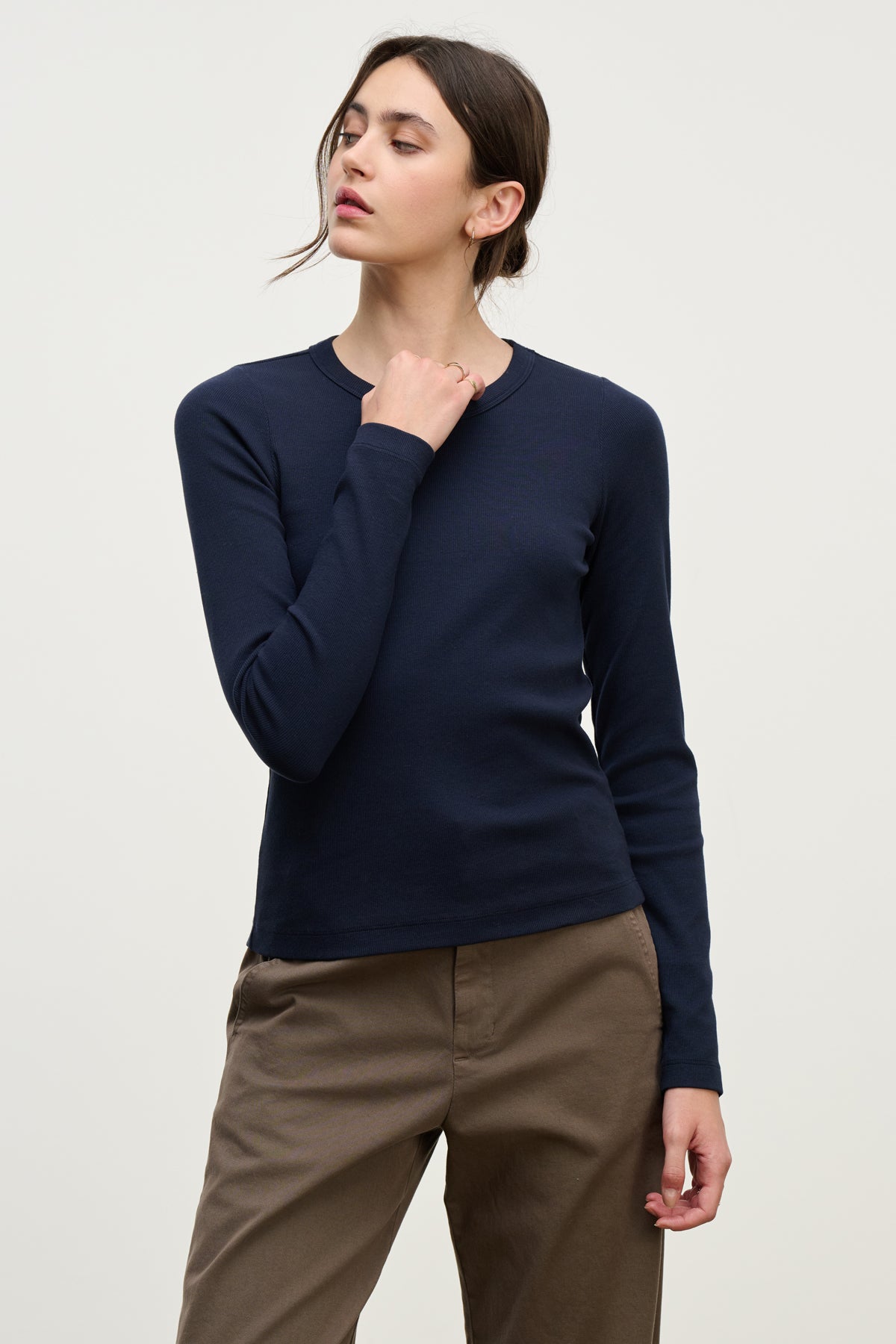 A person stands against a plain background wearing the LOTTA TEE by Velvet by Graham & Spencer in navy, paired with brown pants, one hand raised near their neck and head turned slightly to the side.