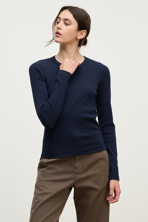 A person stands against a plain background wearing the LOTTA TEE by Velvet by Graham & Spencer in navy, paired with brown pants, one hand raised near their neck and head turned slightly to the side.