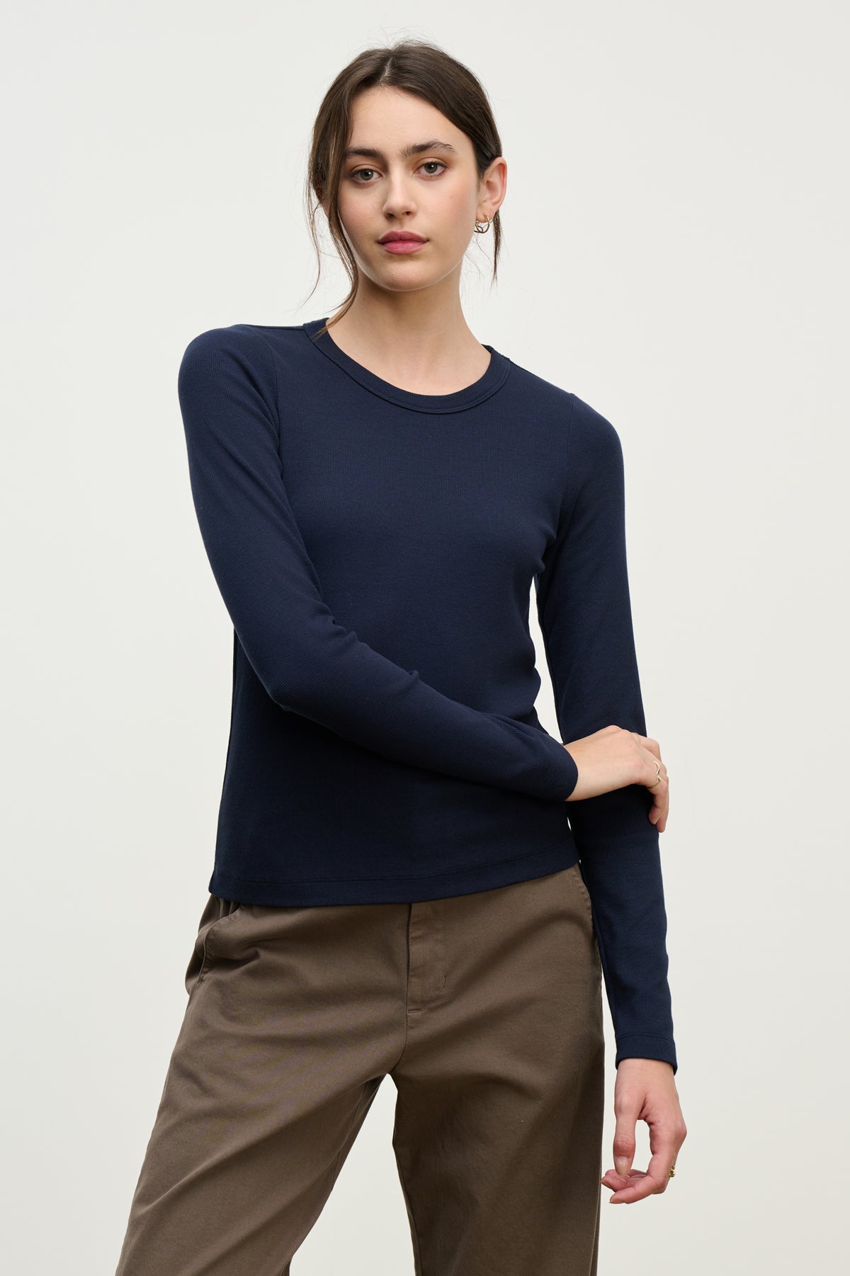 A woman wears the LOTTA TEE by Velvet by Graham & Spencer in navy, paired with brown pants, standing against a plain light background with one arm bent and her hand resting near her elbow.-39814898516161