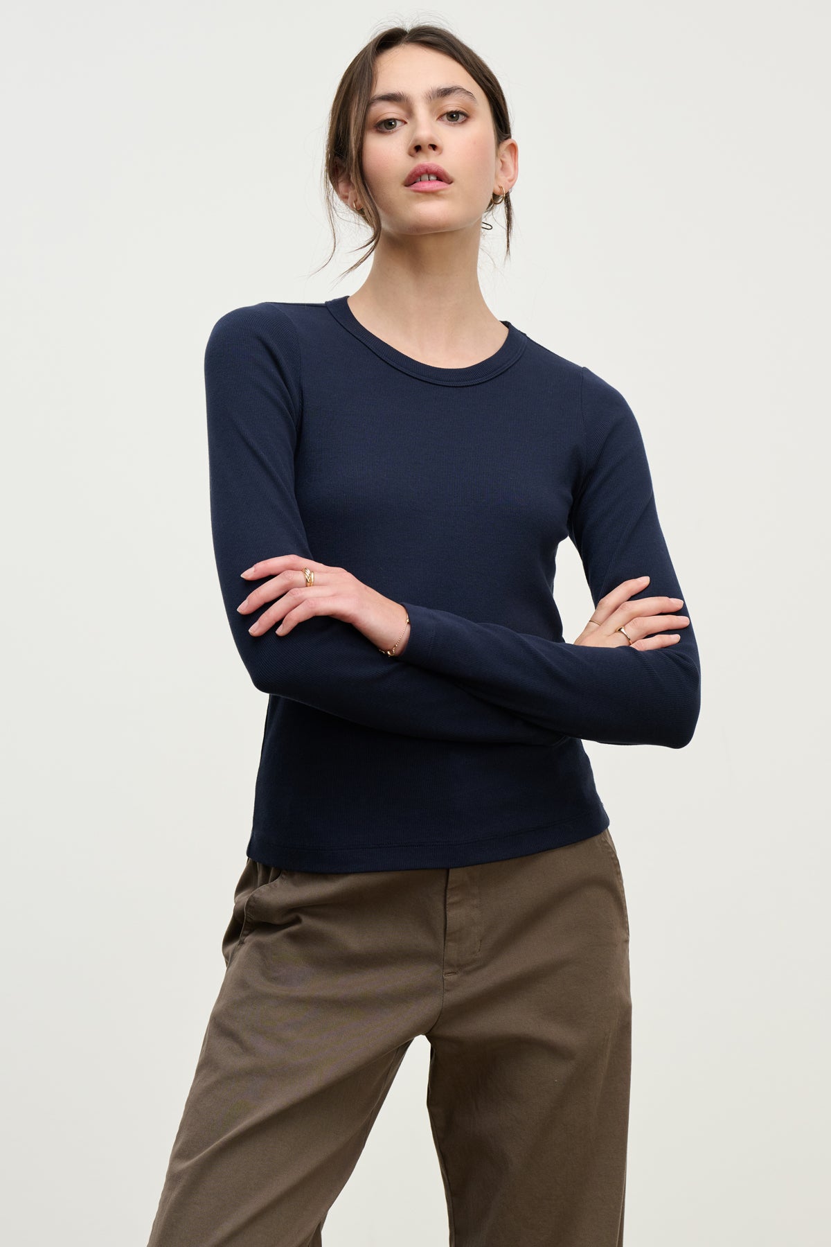 A woman stands with arms crossed, wearing the LOTTA TEE by Velvet by Graham & Spencer—a navy cropped crew neck long sleeve tee—paired with khaki pants against a plain light background.