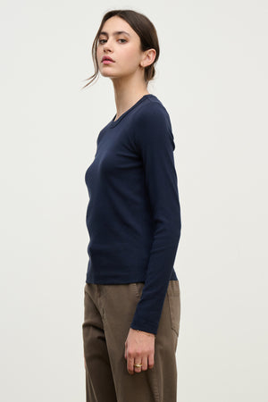 A person wears the LOTTA TEE by Velvet by Graham & Spencer, a long-sleeve ribbed cotton top, with brown pants while standing sideways against a plain light background.