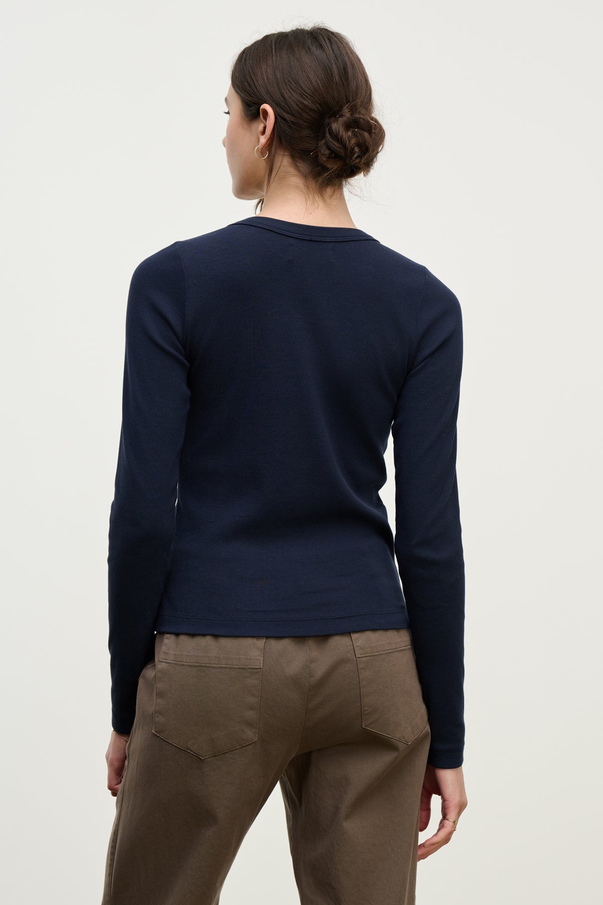 A person with brown hair in a low bun models the LOTTA TEE by Velvet by Graham & Spencer, a navy cropped crew neck long-sleeve top, paired with brown pants and shown from the back against a plain light background.