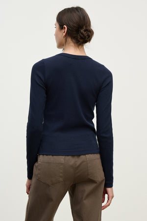 A person with brown hair in a low bun models the LOTTA TEE by Velvet by Graham & Spencer, a navy cropped crew neck long-sleeve top, paired with brown pants and shown from the back against a plain light background.