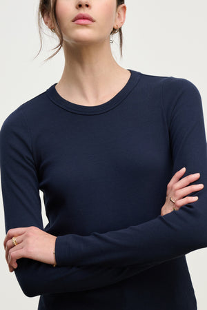 A person with long brown hair, arms crossed and shown from the shoulders down, is wearing the LOTTA TEE by Velvet by Graham & Spencer—a navy blue ribbed cotton long sleeve top.