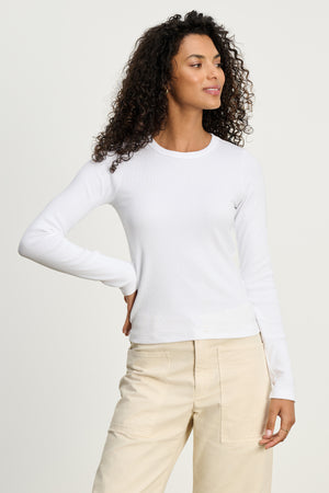 A woman with curly hair wears the LOTTA TEE by Velvet by Graham & Spencer—a fitted ribbed cotton long sleeve top—paired with beige pants, posing with one hand on her hip against a plain light background.