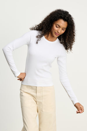 A person models the Velvet by Graham & Spencer LOTTA TEE in white ribbed cotton, paired with light beige pants, standing against a plain light background with one hand on their hip and the other relaxed.