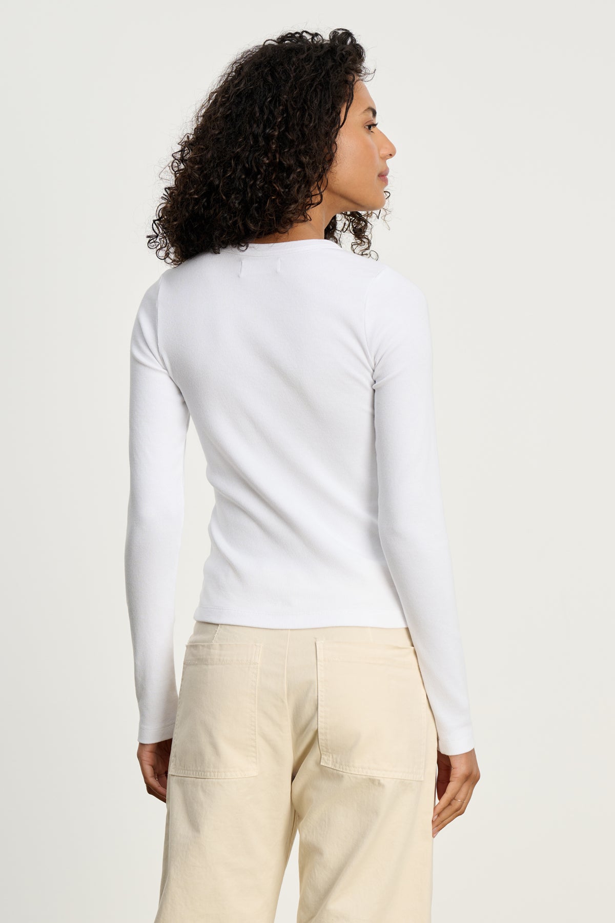 A person with curly hair, seen from behind, wears the LOTTA TEE by Velvet by Graham & Spencer in white and beige pants against a plain light background.-39617521352897
