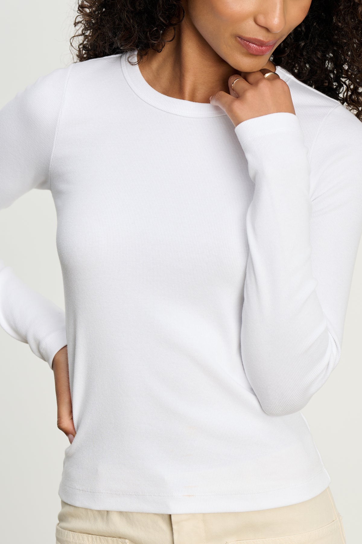 A woman poses with one hand near her chin, wearing the LOTTA TEE by Velvet by Graham & Spencer—a long-sleeve, white ribbed cotton top—paired with light-colored pants.-39617521320129