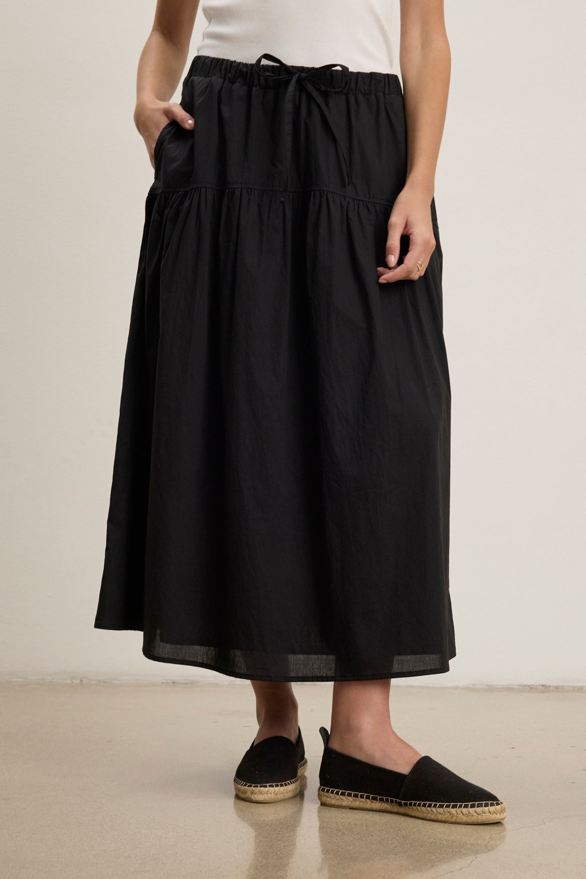 A person wearing the LOUI SKIRT by Velvet by Graham & Spencer—a black, pleated maxi skirt with pockets—paired with a white top and black espadrille flats stands on a light-colored floor.-39123291668673