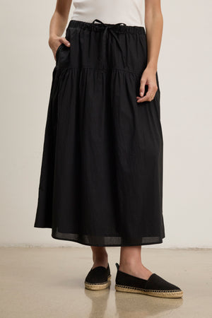 A person wearing the LOUI SKIRT by Velvet by Graham & Spencer—a black, pleated maxi skirt with pockets—paired with a white top and black espadrille flats stands on a light-colored floor.