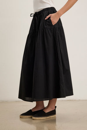 A person wearing the LOUI SKIRT by Velvet by Graham & Spencer—a long black rib poplin blend maxi skirt with pleats and an elastic waistband—paired with black espadrilles on a light-colored floor.