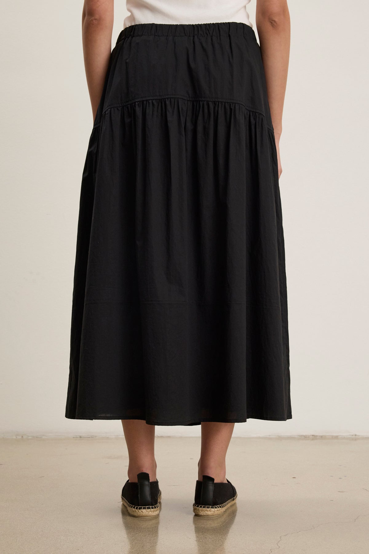 A person, seen from behind against a plain background, wears the LOUI SKIRT by Velvet by Graham & Spencer—a black pleated maxi skirt—paired with black espadrille shoes.-39123291603137
