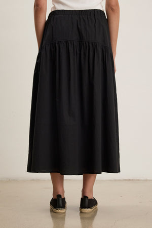 A person, seen from behind against a plain background, wears the LOUI SKIRT by Velvet by Graham & Spencer—a black pleated maxi skirt—paired with black espadrille shoes.