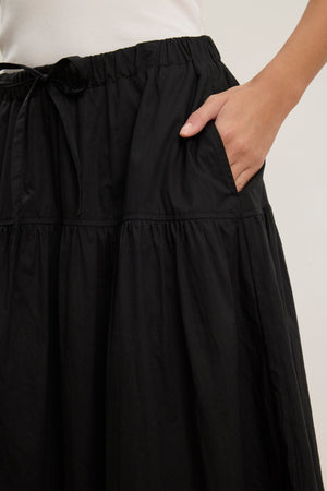 A person wears the LOUI SKIRT by Velvet by Graham & Spencer—a black, pleated maxi skirt featuring an elastic waistband and side pocket for a customizable fit—with one hand in the pocket and a white top.