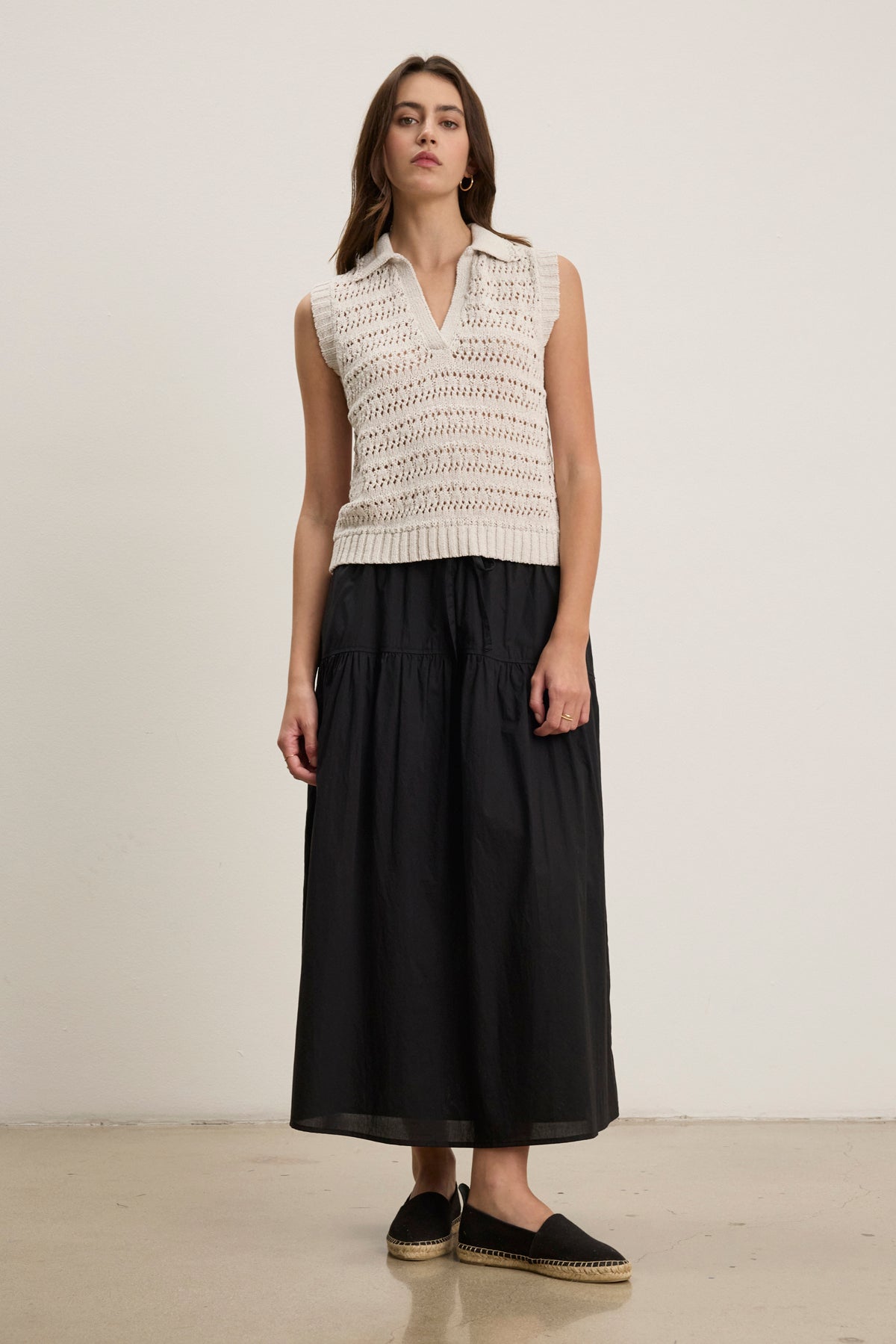 A woman stands against a plain background wearing a sleeveless white knit top, black espadrilles, and the LOUI SKIRT by Velvet by Graham & Spencer.