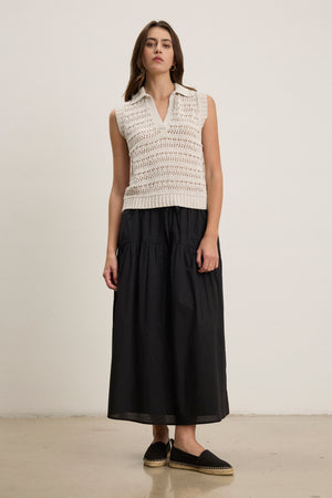 A woman stands against a plain background wearing a sleeveless white knit top, black espadrilles, and the LOUI SKIRT by Velvet by Graham & Spencer.