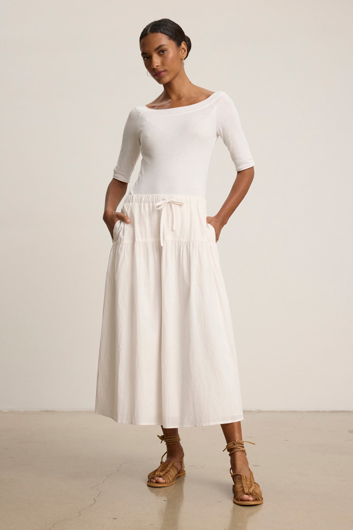 A woman stands indoors in a white top and the LOUI SKIRT by Velvet by Graham & Spencer—a flowing maxi with pleats and pockets—paired with tan lace-up sandals, against a plain background.-39148723536065