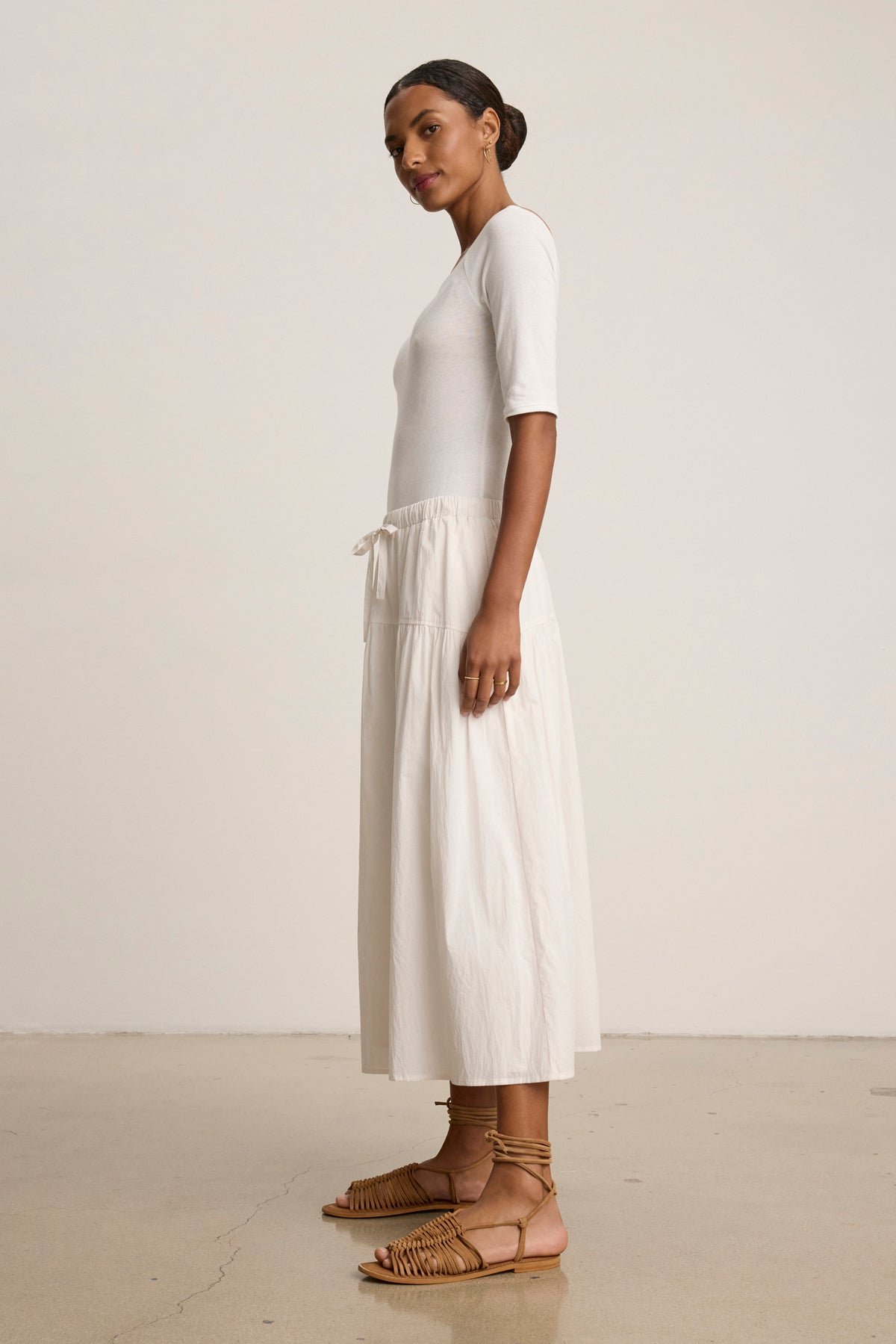 A woman stands sideways against a plain background, wearing the LOUI SKIRT by Velvet by Graham & Spencer—a flowing white maxi with pleated detailing and customizable fit—paired with a white short-sleeve top and tan woven sandals.-39148723503297