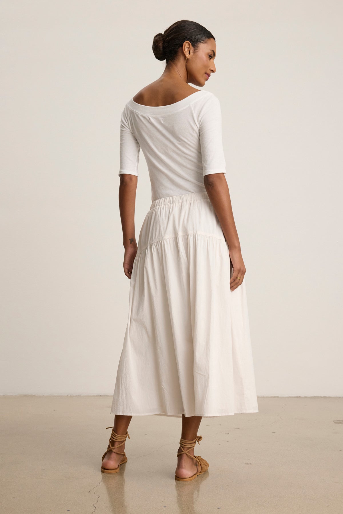 Woman stands on a polished floor, facing away, wearing a white off-the-shoulder rib poplin top, the LOUI SKIRT by Velvet by Graham & Spencer in white maxi style, and brown sandals.-39148723470529