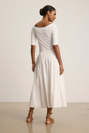 Woman stands on a polished floor, facing away, wearing a white off-the-shoulder rib poplin top, the LOUI SKIRT by Velvet by Graham & Spencer in white maxi style, and brown sandals.