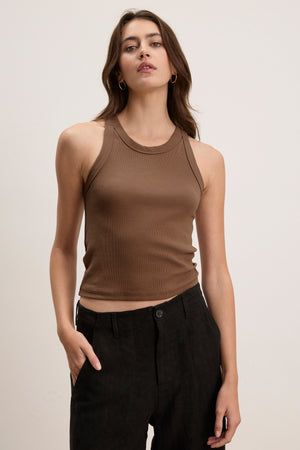 A woman wears the SARI TANK TOP by Velvet by Graham & Spencer—sleeveless and ribbed in brown—with black pants, posing against a plain light background. A modern essential for any wardrobe.