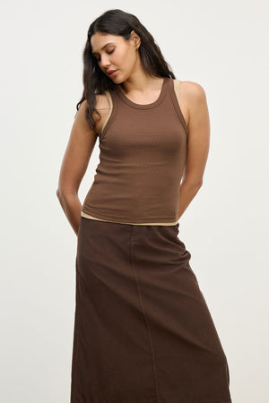 A woman wears the SARI TANK TOP by Velvet by Graham & Spencer with a long brown skirt, standing against a plain light background and looking down with her hands behind her back.