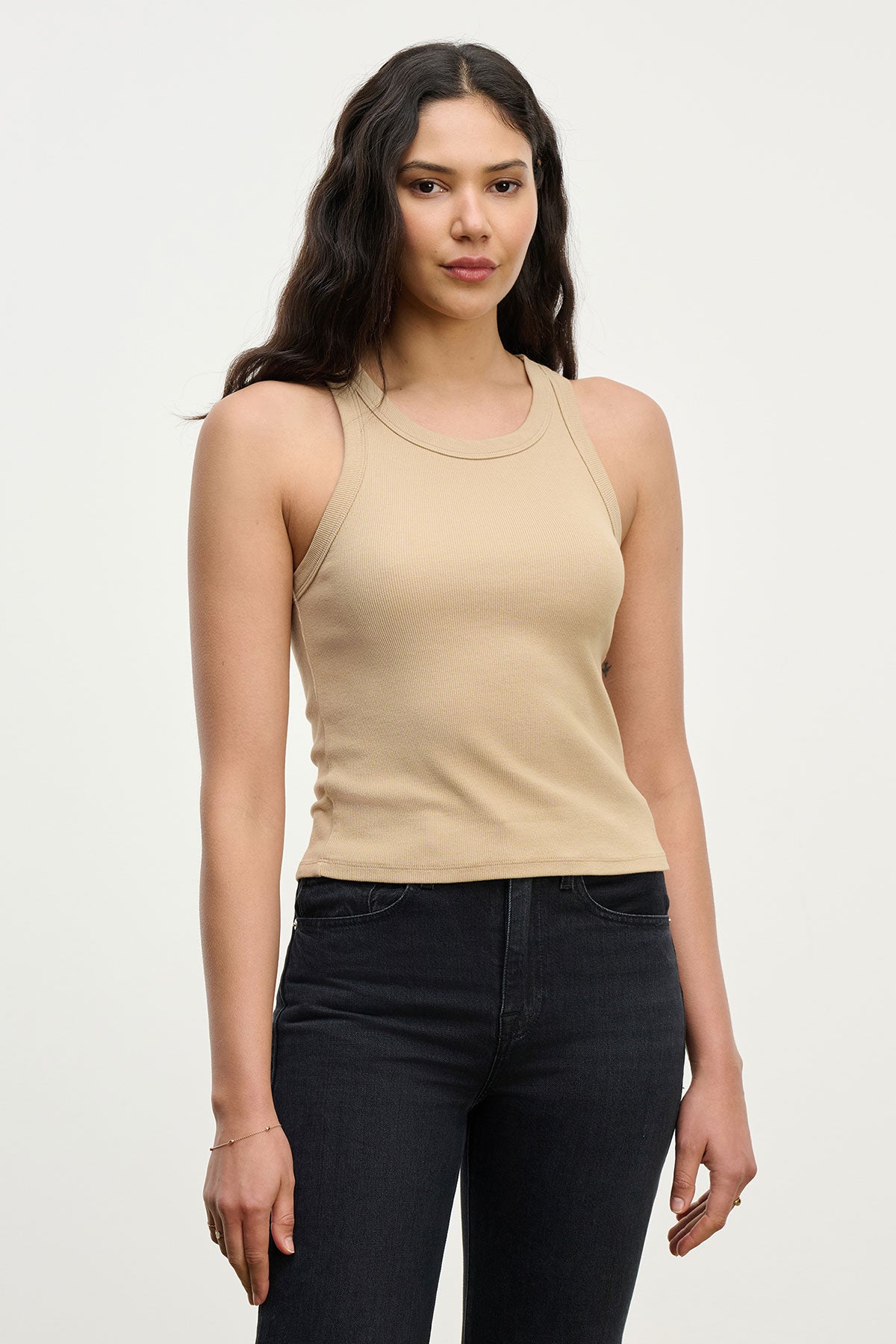 Woman with long dark hair wears the SARI TANK TOP by Velvet by Graham & Spencer in beige, paired with black jeans, standing against a plain light background for a modern essential look.