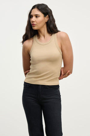 A woman with long dark hair wears the SARI TANK TOP by Velvet by Graham & Spencer in beige, paired with black jeans, standing against a plain light background.